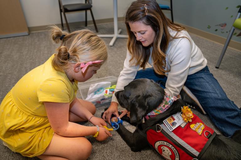 Child life specialist in a clinical room demonstrates toy medical equipment on her facility dog for a child
