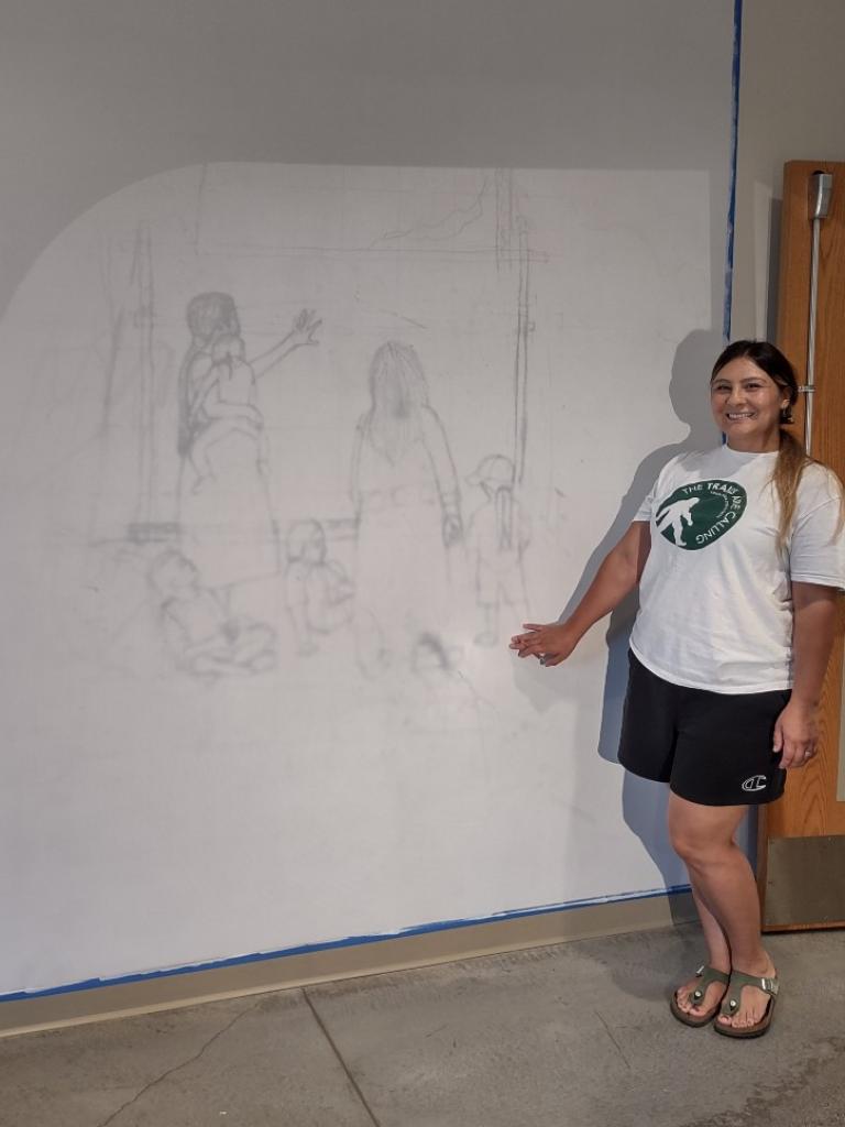 Wóokiye Wiŋ, an Indigenous woman with a long ponytail, a big smile, and a white-and-green tee shirt points to a wall where a drawing of two adults and four children is projected