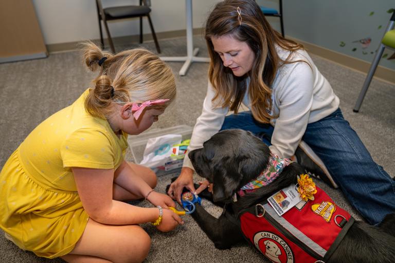 Child life specialist in a clinical room demonstrates toy medical equipment on her facility dog for a child