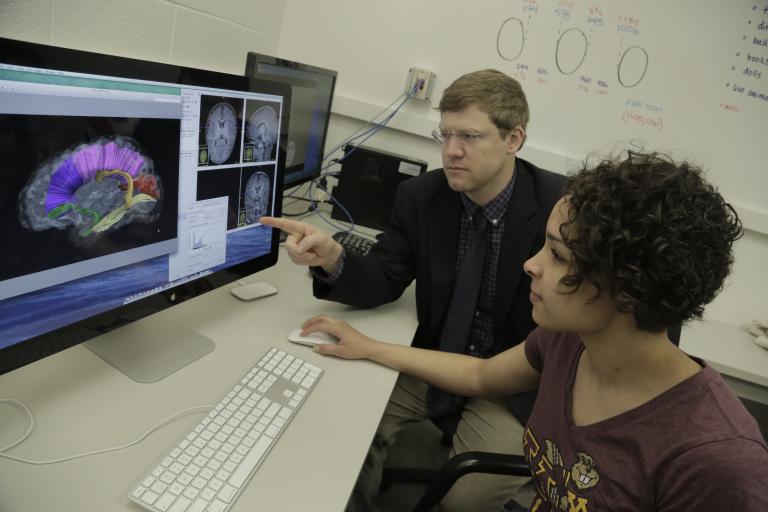 A professor sits with a student at a desk pointing at an image of a brain on a screen