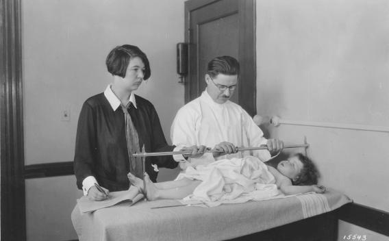 1920s era photo of a researcher measuring a young child with an assistant standing off to the side