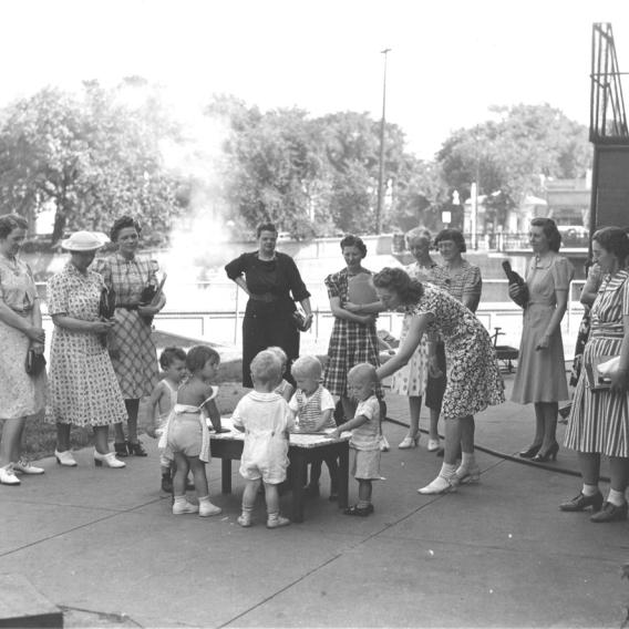 1940s era photo of a group of women standing outside around a small group of toddlers playing at a table