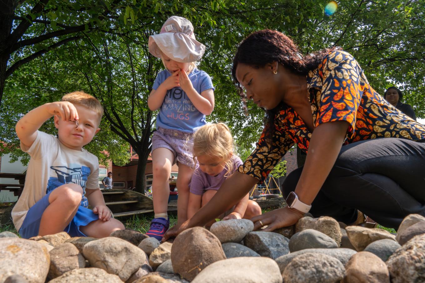 A preschool teacher plays outside with a group of preschoolers in a bed of rocks