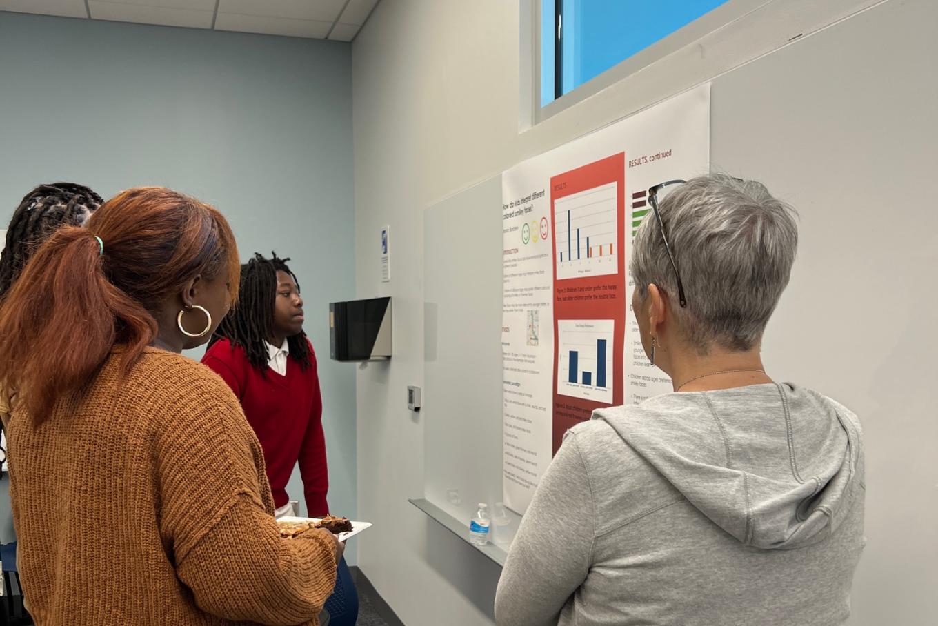 Middle school student stands in front of a research poster presenting to three adults