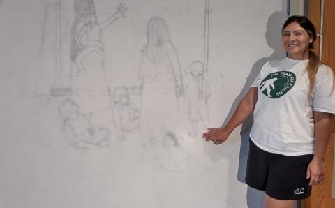 Wóokiye Wiŋ, an Indigenous woman with a long ponytail, a big smile, and a white-and-green tee shirt points to a wall where a drawing of two adults and four children is projected